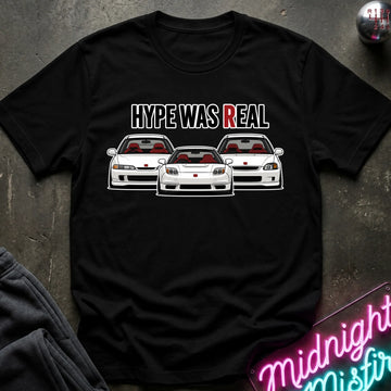 A black short-sleeve graphic t-shirt laid flat on a dark textured surface. The shirt features a white and red graphic print of three sports cars with the text 'HYPE WAS REAL' above them, with 'R' highlighted in red.