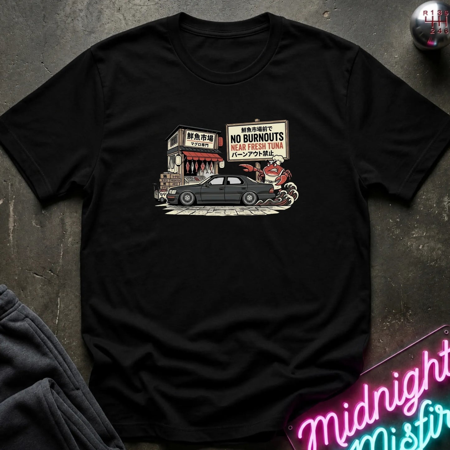 A black short-sleeve graphic t-shirt laid flat on a dark textured surface. The graphic features a vintage-style illustration of a black car parked in front of a shop with a sign that reads 'NO BURNOUTS NEAR FRESH TUNA'.