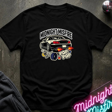 A black short-sleeve graphic t-shirt laid flat on a dark textured surface. The shirt features a graphic of a black sports car with flames, a blue tire, and the text 'MIDNIGHT MISFIRE' and 'POP POP'.