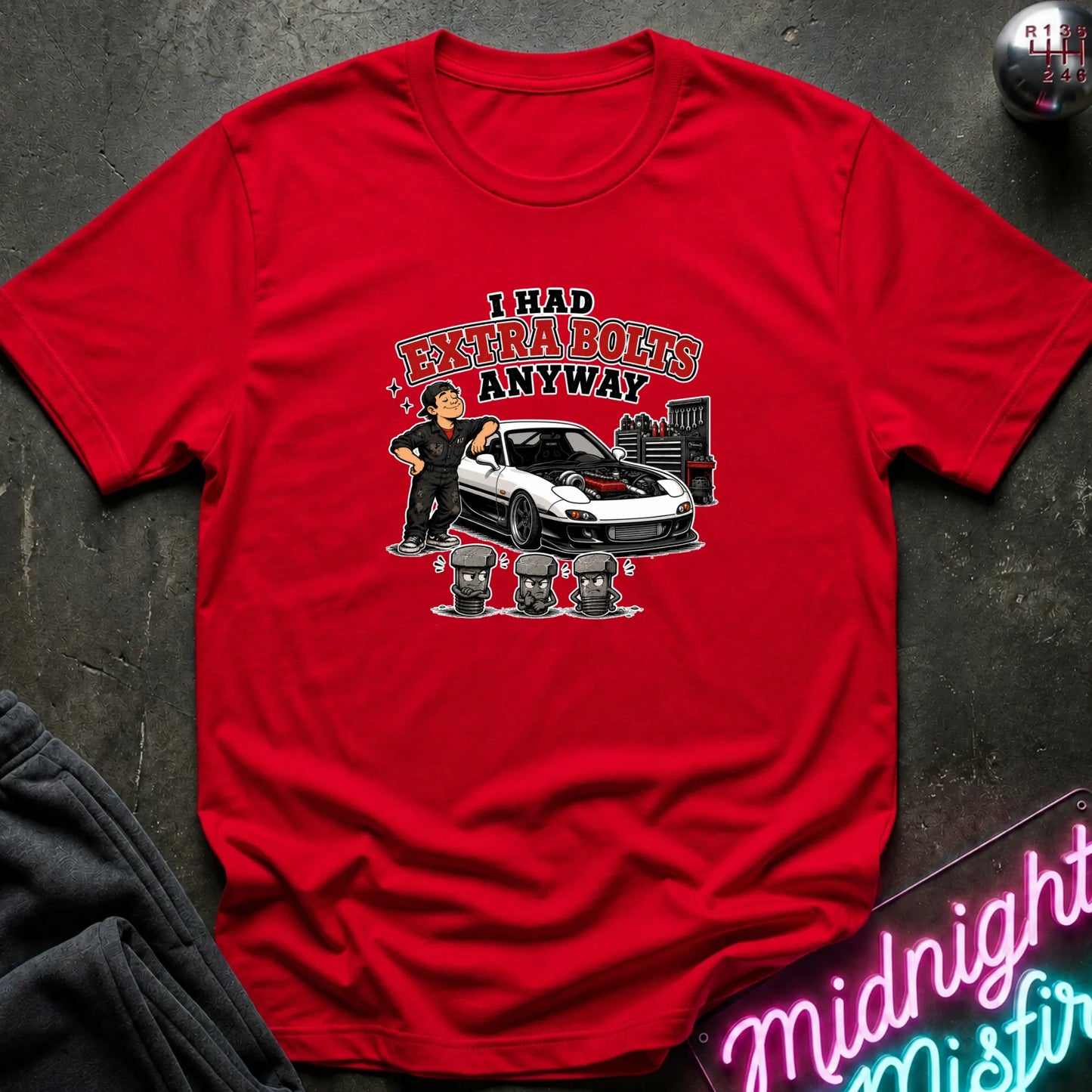 A red short-sleeve graphic t-shirt laid flat on a dark textured surface. The graphic features a cartoon of a man standing next to a white sports car with red detailing, with the text 'I HAD EXTRA BOLTS ANYWAY' in red and black.