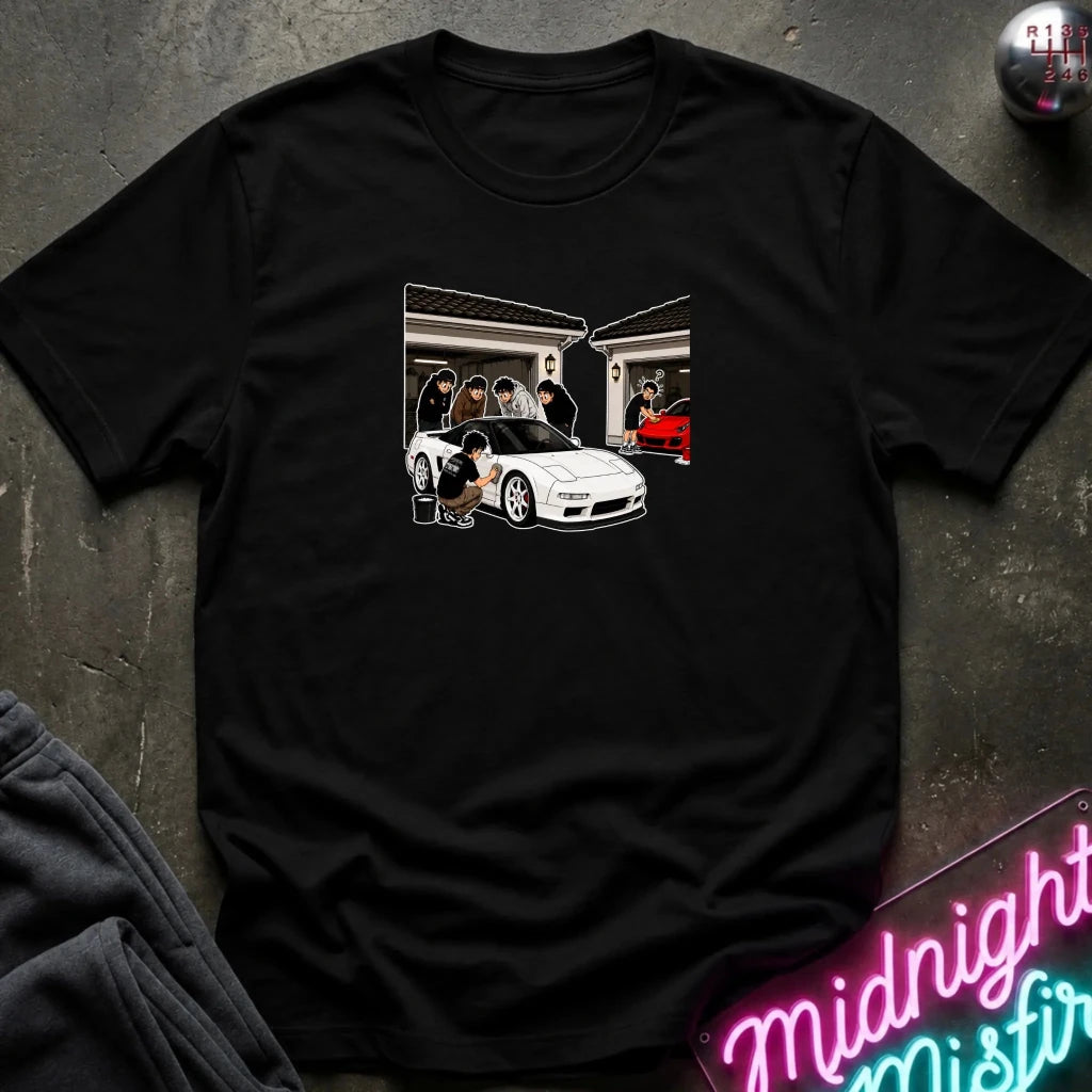A black short-sleeve graphic t-shirt laid flat on a dark textured surface. The graphic shows a white sports car in front of a house with several figures.