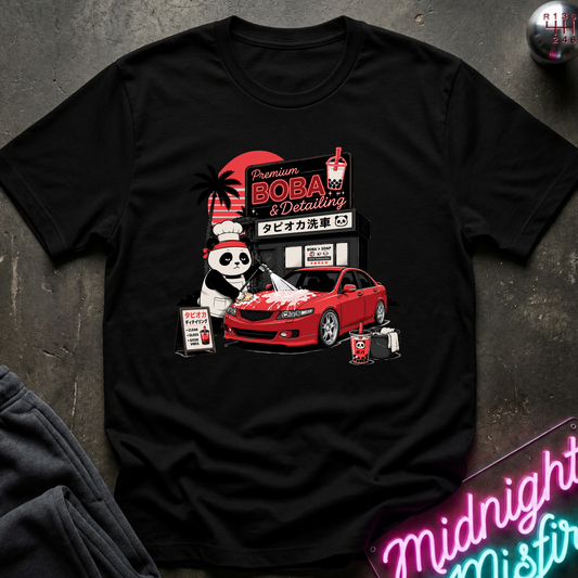A black short-sleeve graphic T-shirt laid flat on a dark textured surface. The shirt features a colorful retro-style print with a panda, a red car, a storefront sign reading 'Premium BOBA & Detailing,' and small drink cups.