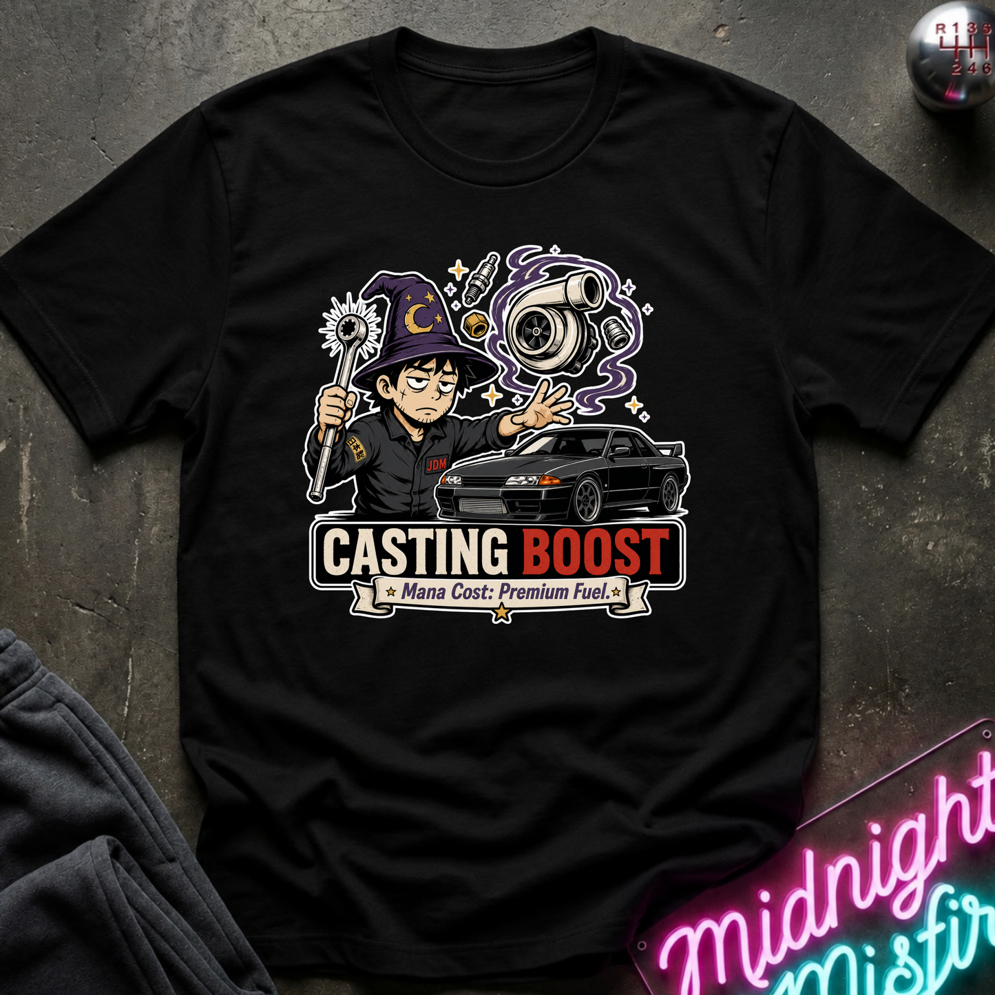 A black short-sleeve graphic T-shirt laid flat on a dark textured surface. The shirt features a colorful cartoon wizard holding a staff next to a black sports car, with the text 'CASTING BOOST' and 'Mana Cost: Premium Fuel.