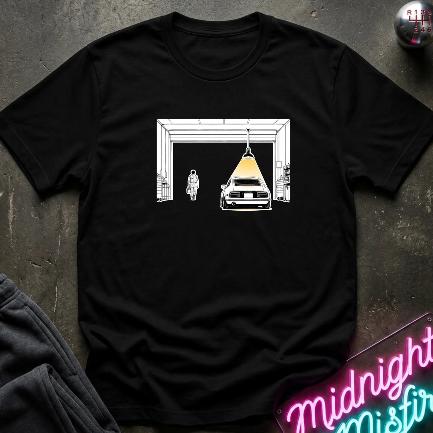 A black short-sleeve t-shirt laid flat on a dark textured surface, featuring a graphic print of a car under a hanging lamp inside a garage structure. 