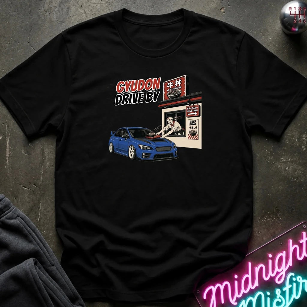 A black short-sleeve graphic t-shirt laid flat on a dark textured surface. The graphic features a blue sports car in front of a Japanese-style shop with the text 'GYUDON DRIVE BY'.