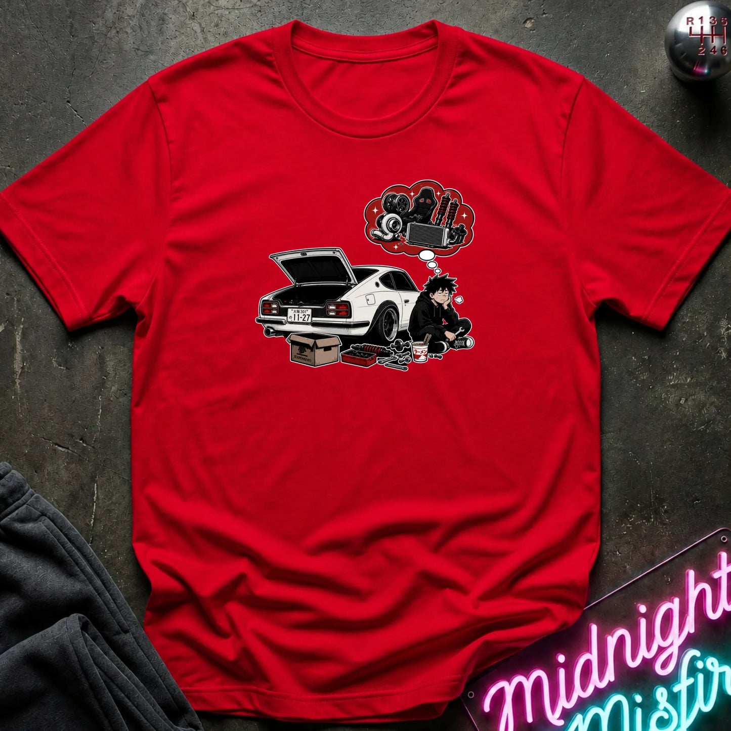 A red short-sleeve graphic t-shirt laid flat on a dark textured surface. The graphic features a white sports car with its trunk open, a cartoon character sitting beside it, and a red thought bubble filled with car parts.