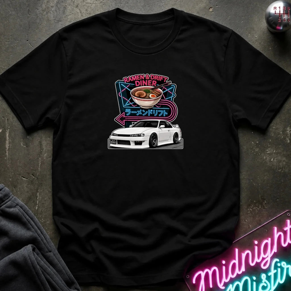 A black short-sleeve graphic t-shirt laid flat on a dark textured surface. The graphic features a white sports car beneath a ramen bowl with pink and blue neon-style text reading 'Ramen & Drift Diner' and Japanese characters.