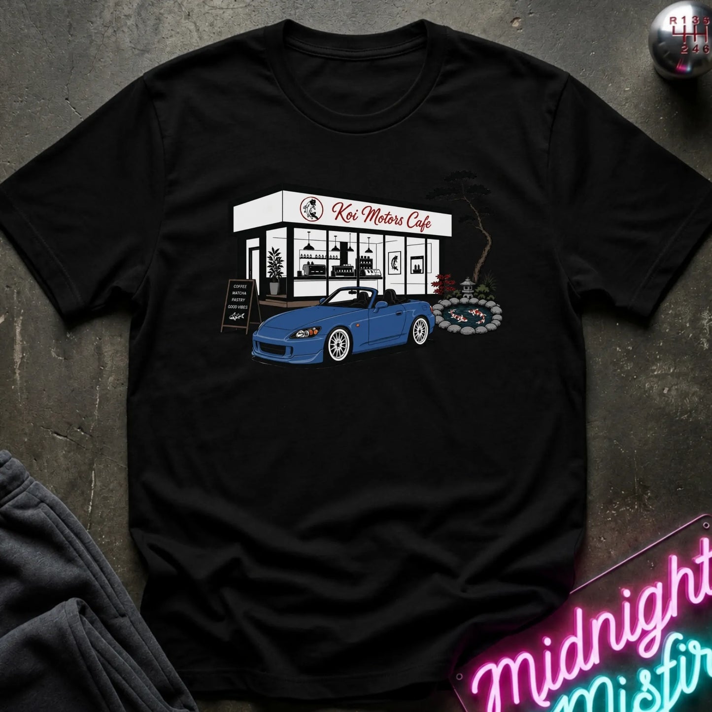 A black short-sleeve t-shirt laid flat on a dark textured surface, featuring a graphic print of a blue convertible sports car parked in front of a white cafe building labeled 'Koi Motor Cafe'.