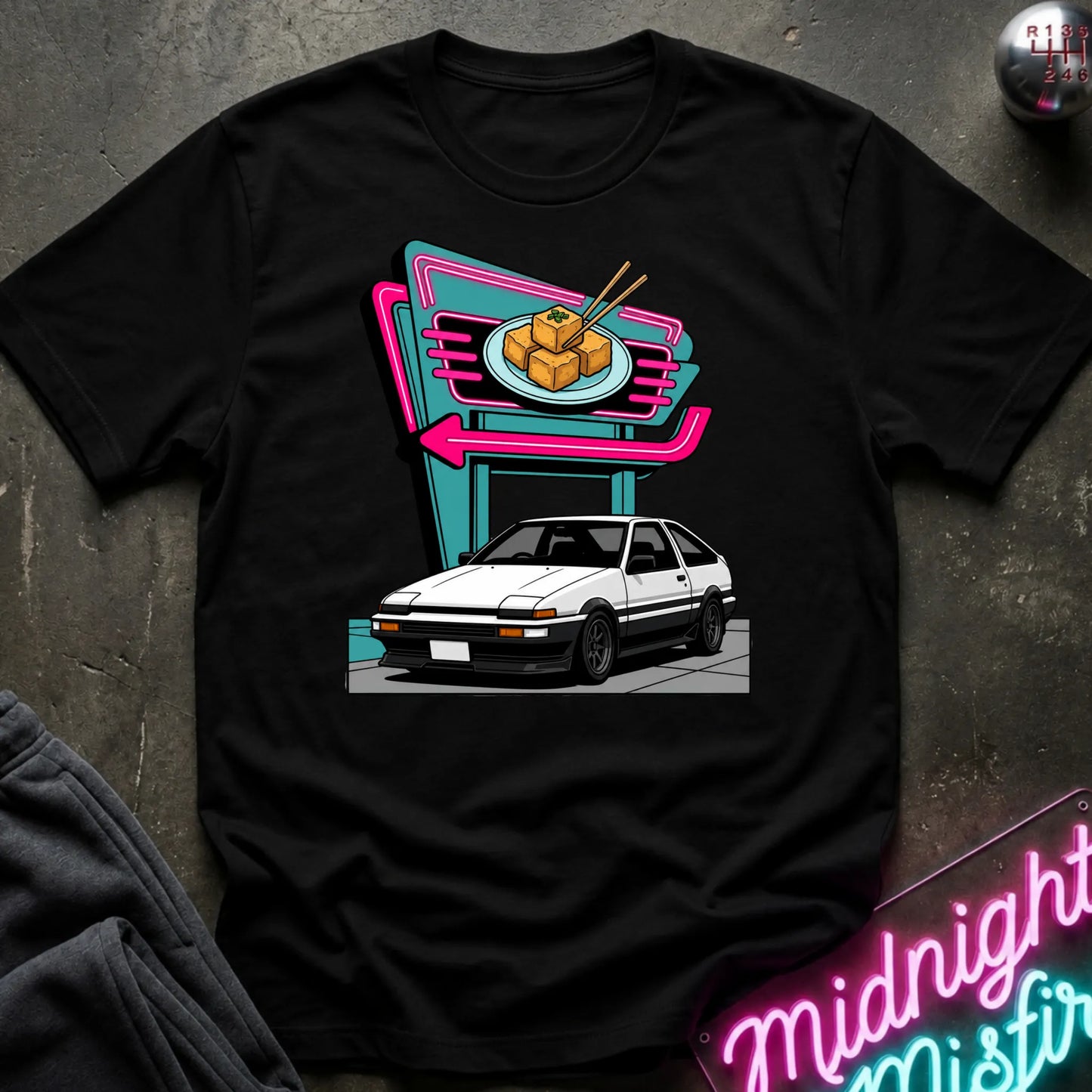 A black short-sleeve crewneck t-shirt featuring a retro-style graphic of a white sports car parked in front of a neon sign. The shirt is displayed flat on a dark textured surface with a dark sweatshirt and a neon sign partially visible as props.