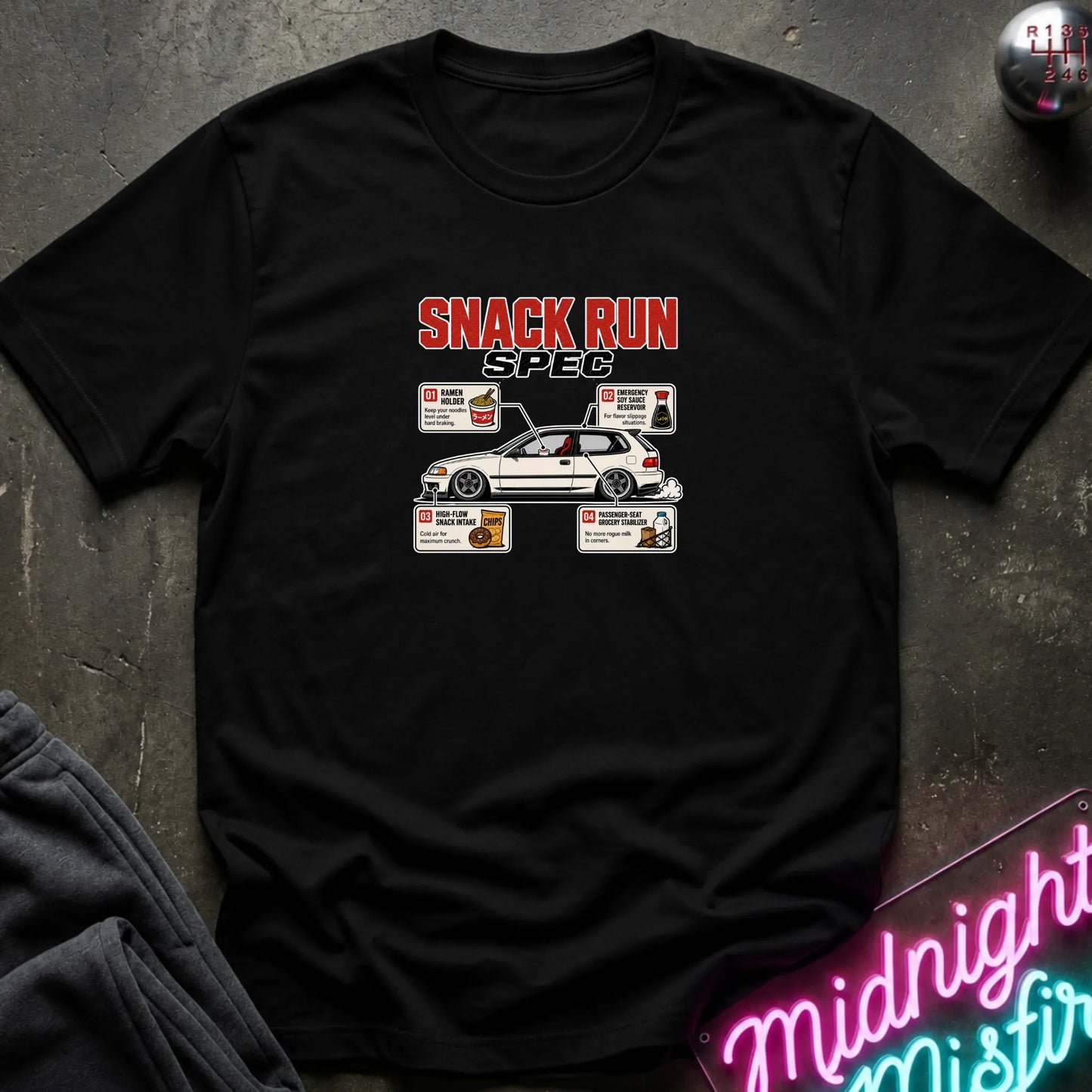 A black short-sleeve t-shirt laid flat on a dark textured surface, featuring a 'SNACK RUN SPEC' graphic with a white car and small food illustrations. A neon sign reading 'Midnight Misfire' is partially visible in the bottom right corner.