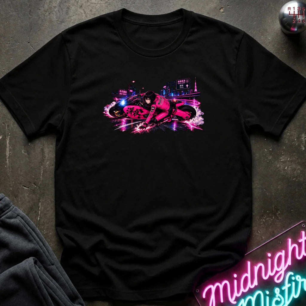 A black short-sleeve crewneck t-shirt laid flat on a dark textured surface, featuring a vibrant neon pink and purple graphic of a motorcycle with Japanese text on the chest. A neon sign and folded dark fabric are partially visible in the corners.