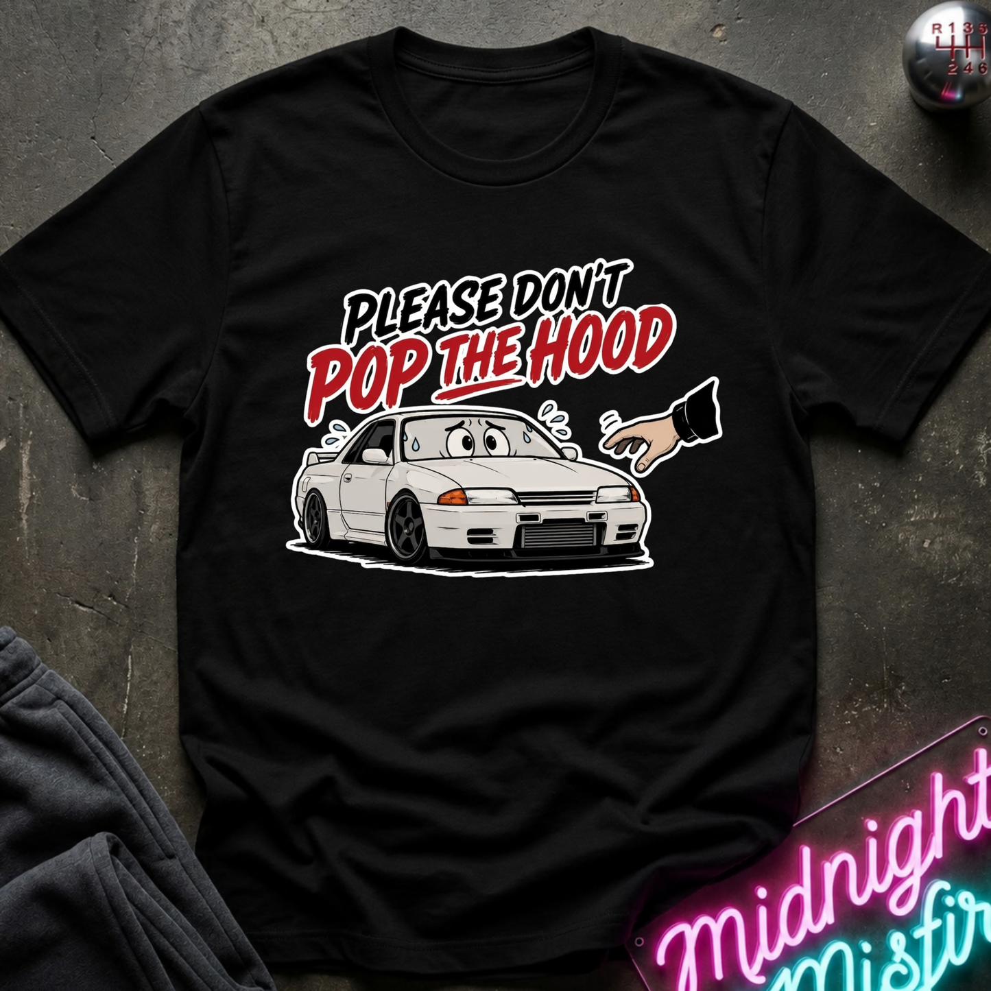 A black short-sleeve graphic T-shirt laid flat on a dark textured surface. The shirt features a cartoon white sports car with a surprised face and the text 'PLEASE DON'T POP THE HOOD' in white and red.