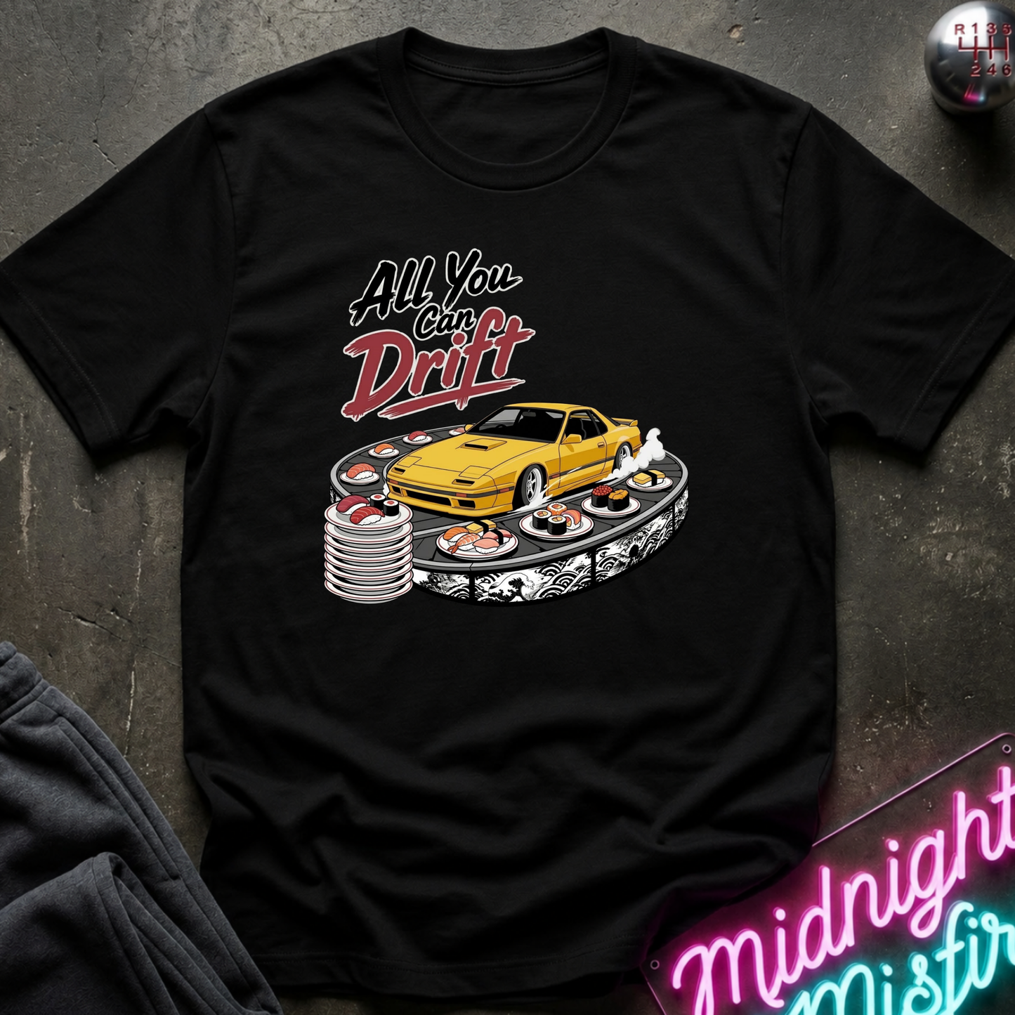 A black short-sleeve graphic T-shirt laid flat on a dark textured surface. The shirt features a printed design of a yellow sports car on a drift track with sushi plates and the text 'All You Can Drift' in stylized white and red lettering.