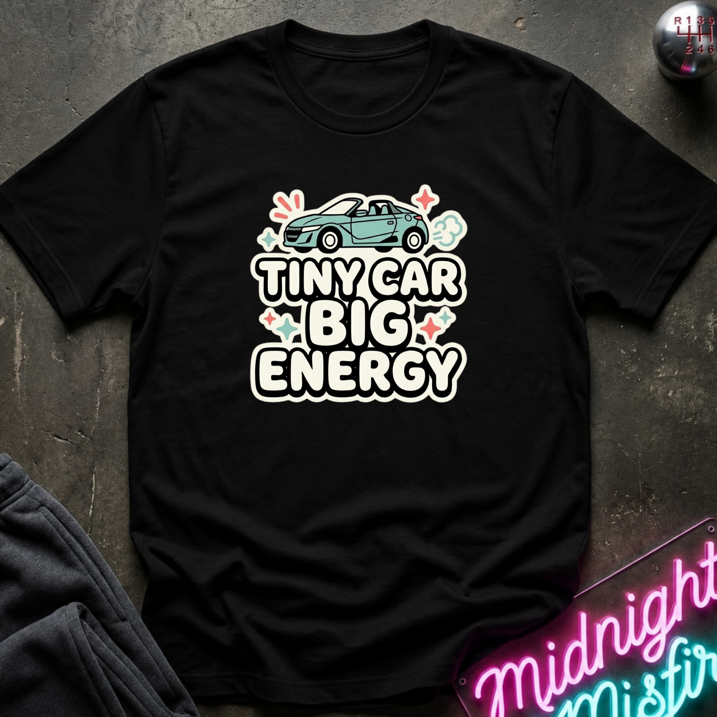 A black short-sleeve graphic t-shirt laid flat on a dark textured surface. The shirt features a graphic of a small teal convertible car above the text 'TINY CAR BIG ENERGY' in white outlined letters with small pink and green floral accents.