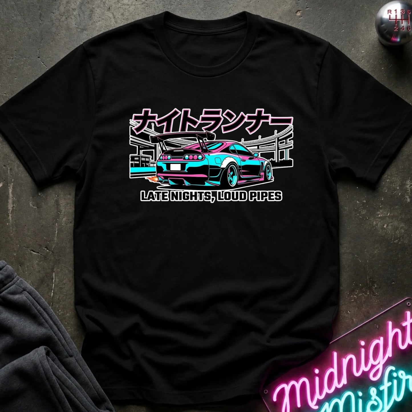 A black short-sleeve graphic T-shirt laid flat on a dark textured surface. The shirt features a colorful print of a sports car with Japanese text above and the phrase 'LATE NIGHTS, LOUD PIPES' below.
