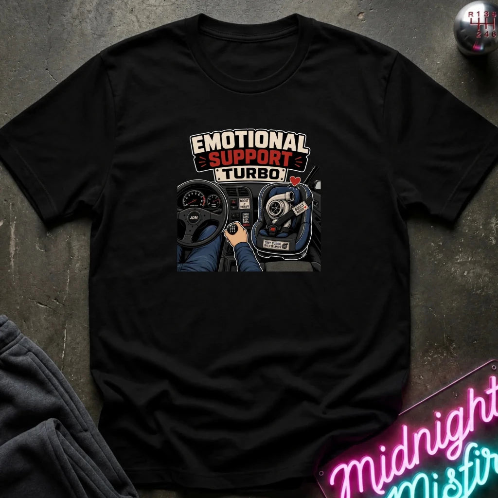 A black short-sleeve graphic t-shirt laid flat on a dark textured surface. The graphic features the words 'EMOTIONAL SUPPORT TURBO' above an illustration of a hand on a steering wheel and gear shift console.