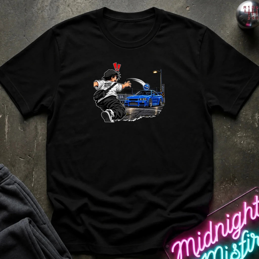 A black short-sleeve crewneck t-shirt laid flat on a dark textured surface. The center of the shirt features a graphic print of an anime character in a black and white outfit with a hat, next to a blue sports car under a streetlamp.