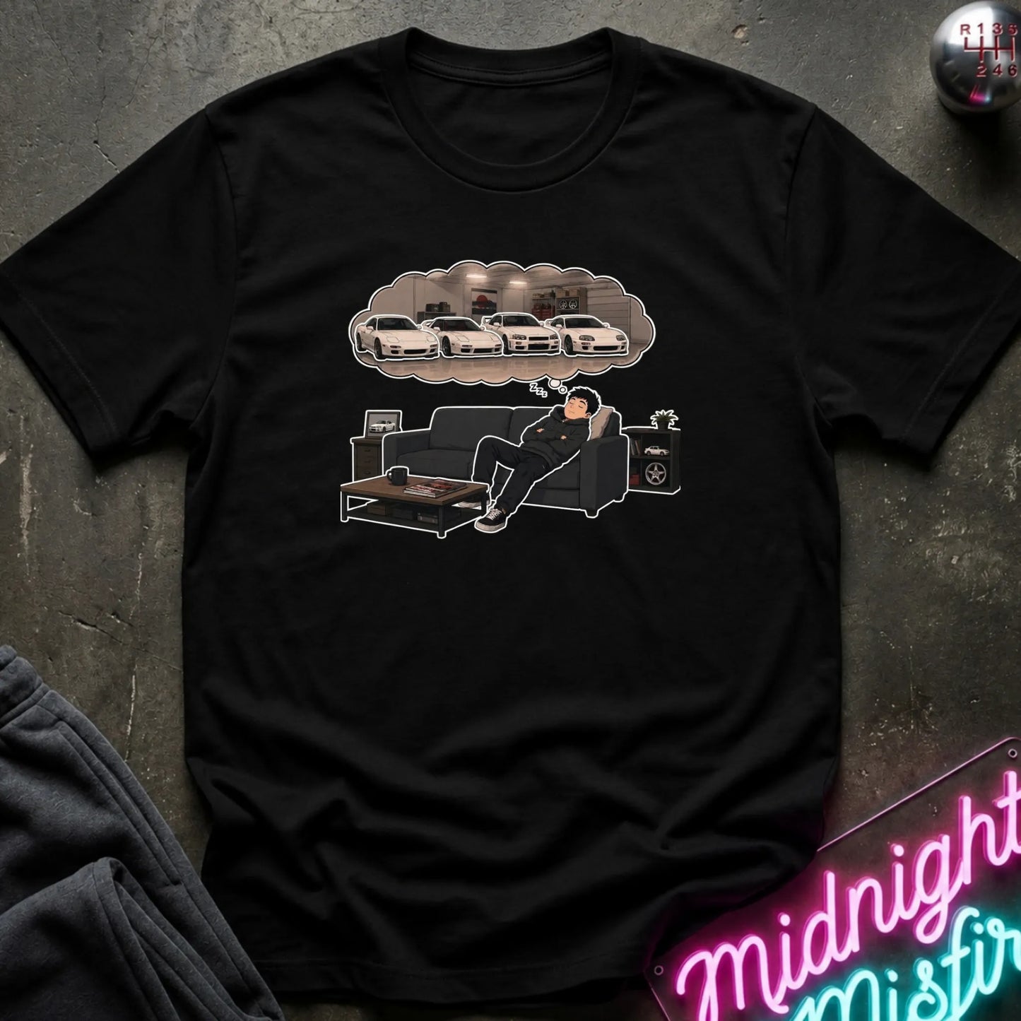 A black short-sleeve graphic t-shirt laid flat on a dark textured surface. The graphic shows a cartoon figure lounging on a couch beneath a thought bubble containing white cars.