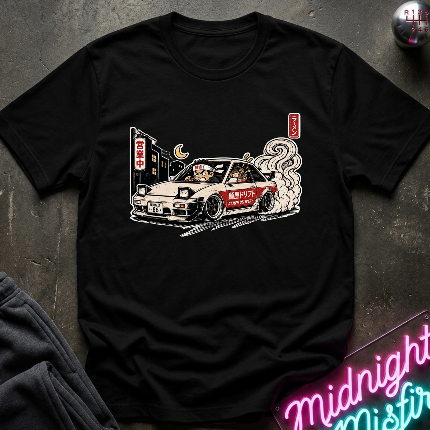 A black short-sleeve graphic t-shirt laid flat on a dark textured surface. The shirt features a detailed illustration of a white and red sports car with smoke, a crescent moon, and Japanese-style signs.