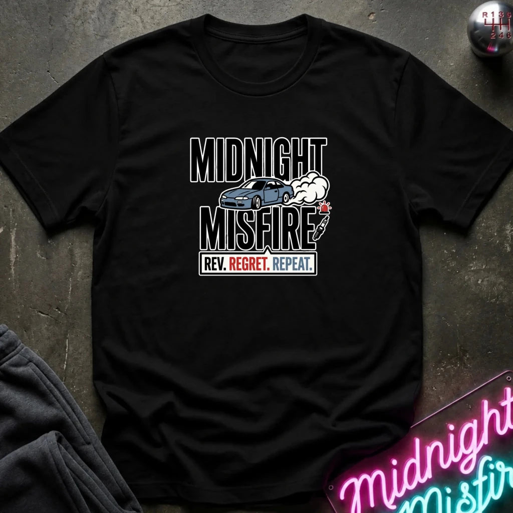 A black short-sleeve graphic T-shirt laid flat on a dark textured surface. The shirt features a distressed white and blue graphic with the text 'MIDNIGHT MISFIRE' and 'REV.