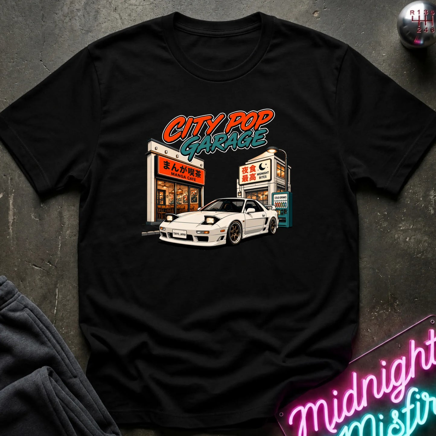 A black short-sleeve graphic t-shirt laid flat on a dark textured surface. The graphic features a white sports car parked in front of a cafe with the text 'CITY POP GARAGE' in orange and teal.