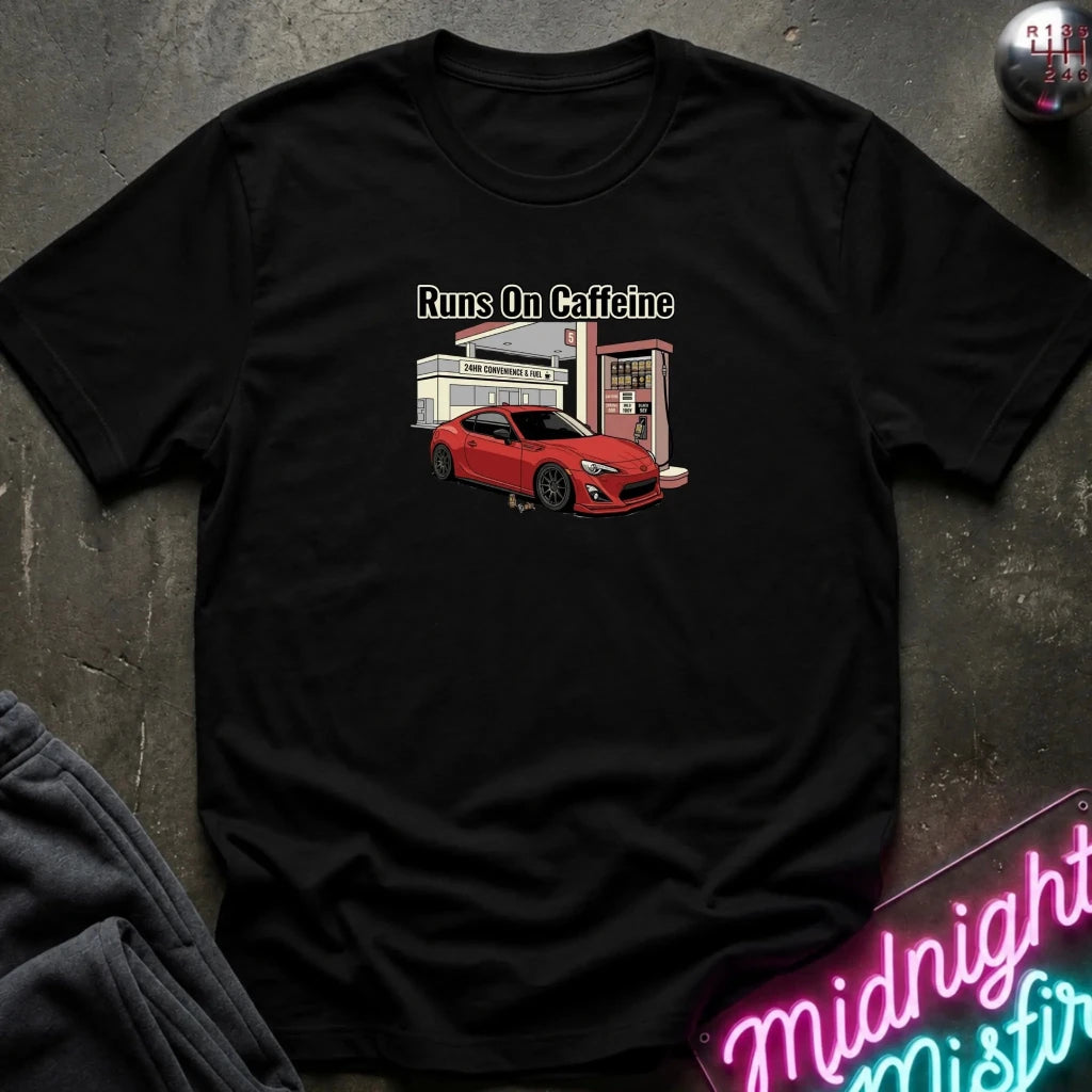 A black short-sleeve graphic t-shirt laid flat on a dark textured surface. The shirt features a graphic of a red sports car at a gas station with the text 'Runs On Caffeine' above it.