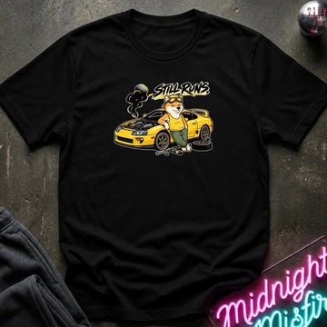 A black short-sleeve graphic t-shirt laid flat on a dark textured surface. The graphic features a cartoon dog in a mechanic hat standing next to a yellow sports car with the text 'Still runs' above it.