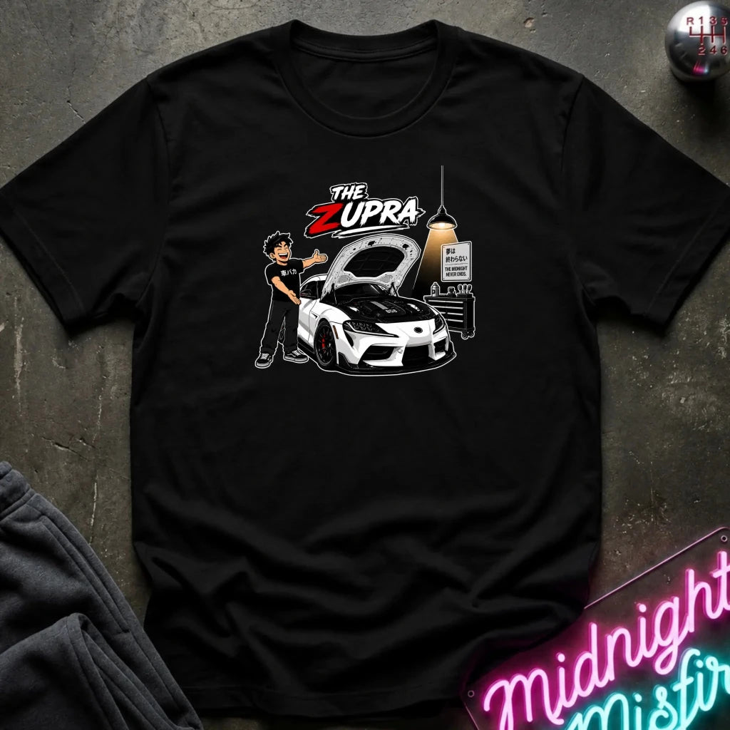 A black short-sleeve graphic t-shirt laid flat on a dark textured surface. The graphic features a cartoon illustration of a man standing next to a white sports car with its hood open, under the text 'THE ZUPRA'.