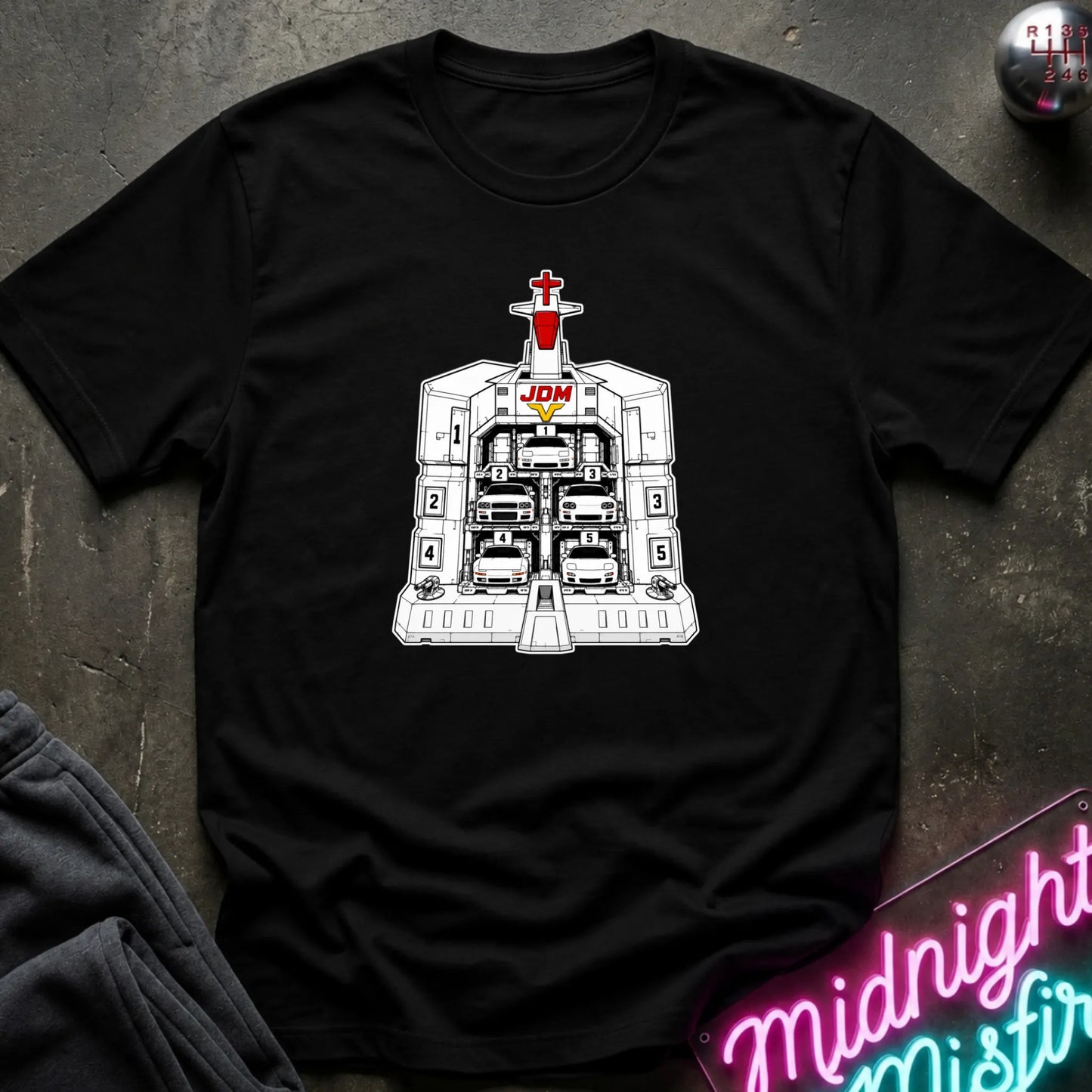 A black short-sleeve crewneck t-shirt laid flat on a dark textured surface, featuring a black and white graphic of the JDM (Japanese Domestic Market) cars in a launch bay.
