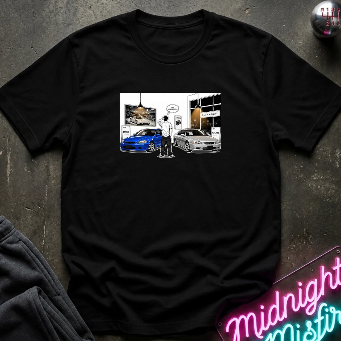 A black short-sleeve crewneck t-shirt laid flat on a dark textured surface. The center of the shirt features a rectangular graphic print depicting a person choosing cars, one blue and one silver.