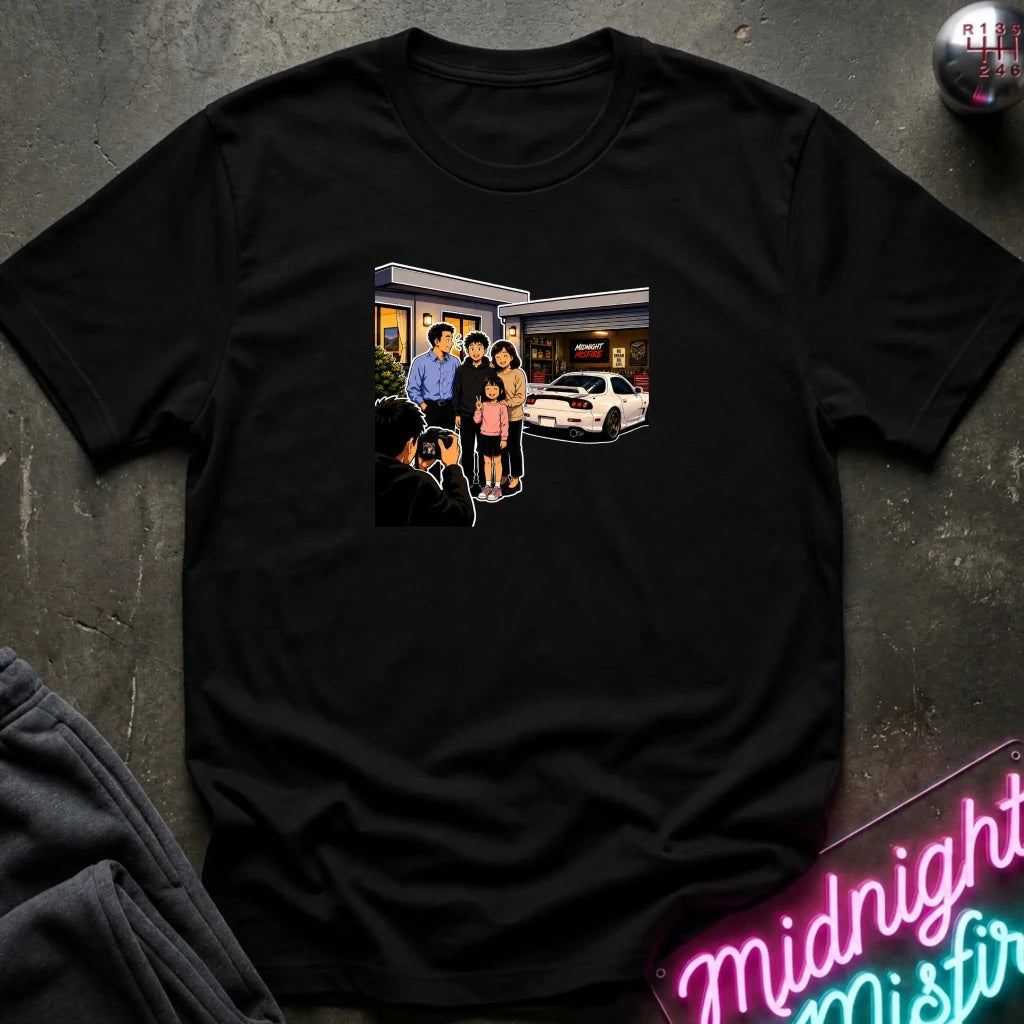 A black short-sleeve crewneck t-shirt laid flat on a dark textured surface, featuring a rectangular graphic print of a family standing outside a building taking a photo.