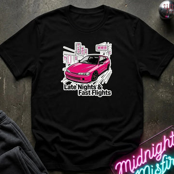 A black short-sleeve graphic t-shirt laid flat on a dark textured surface. The shirt features a graphic of a pink car in front of a white city skyline with the text 'Late Nights & Fast Flights'.