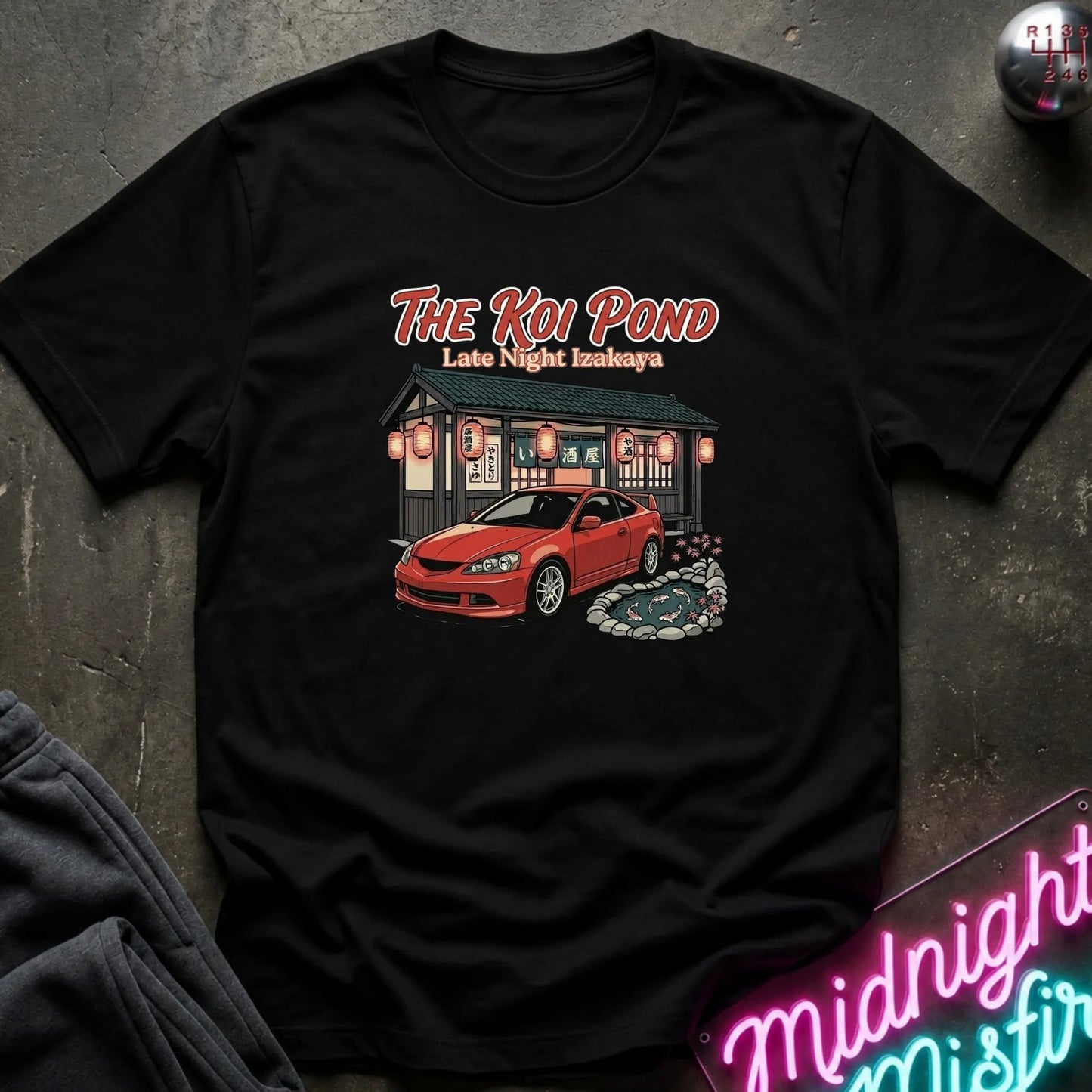 A black short-sleeve graphic t-shirt laid flat on a dark textured surface. The graphic features the text 'THE KOI POND Late Night Hamburger' above an illustration of a red sports car parked in front of a restaurant with red lanterns and a koi pond.