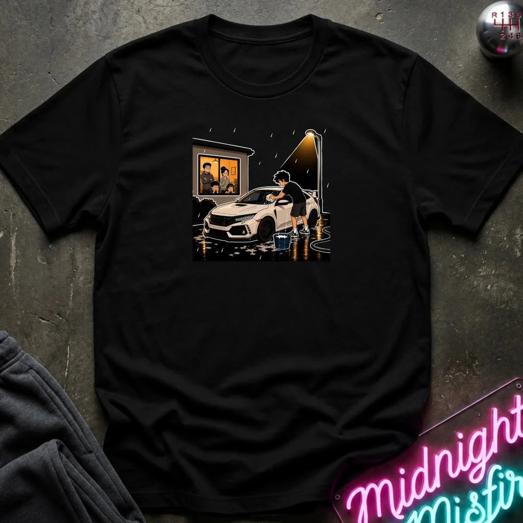 A black short-sleeve graphic t-shirt laid flat on a dark textured surface. The graphic shows a white car in the rain with a person standing beside it and a lit window in the background.
