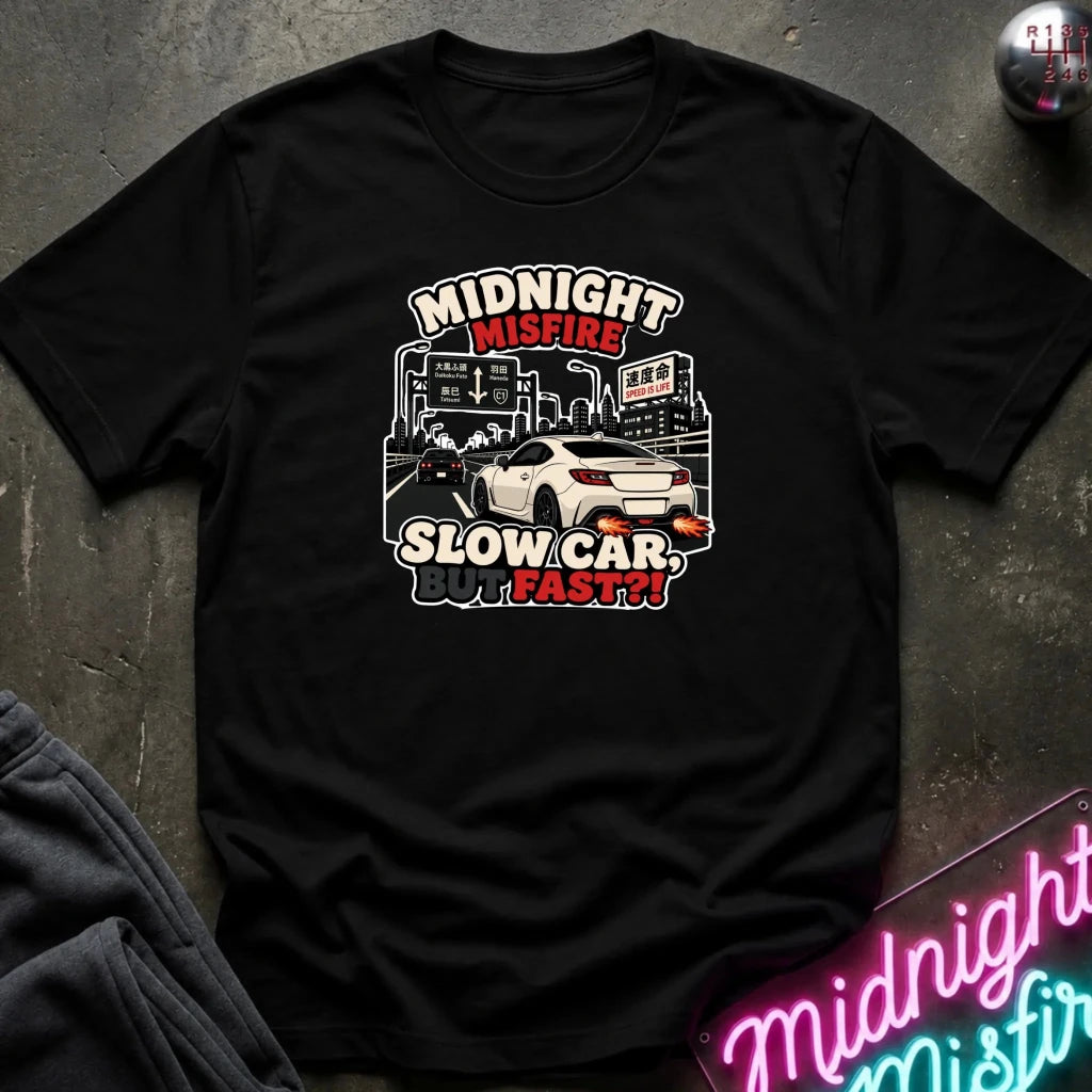A black short-sleeve graphic t-shirt laid flat on a dark textured surface. The graphic features a white sports car and the text 'MIDNIGHT MISFIRE SLOW CAR FAST!!' in stylized red, white, and black lettering.