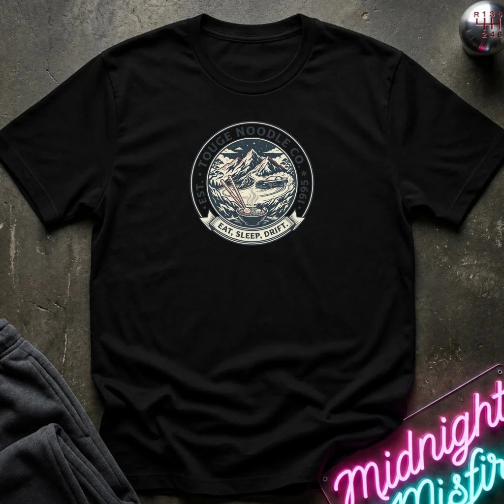 A black short-sleeve t-shirt laid flat on a dark textured surface, featuring a circular vintage-style graphic on the chest with mountains, a sleeping figure, and the text 