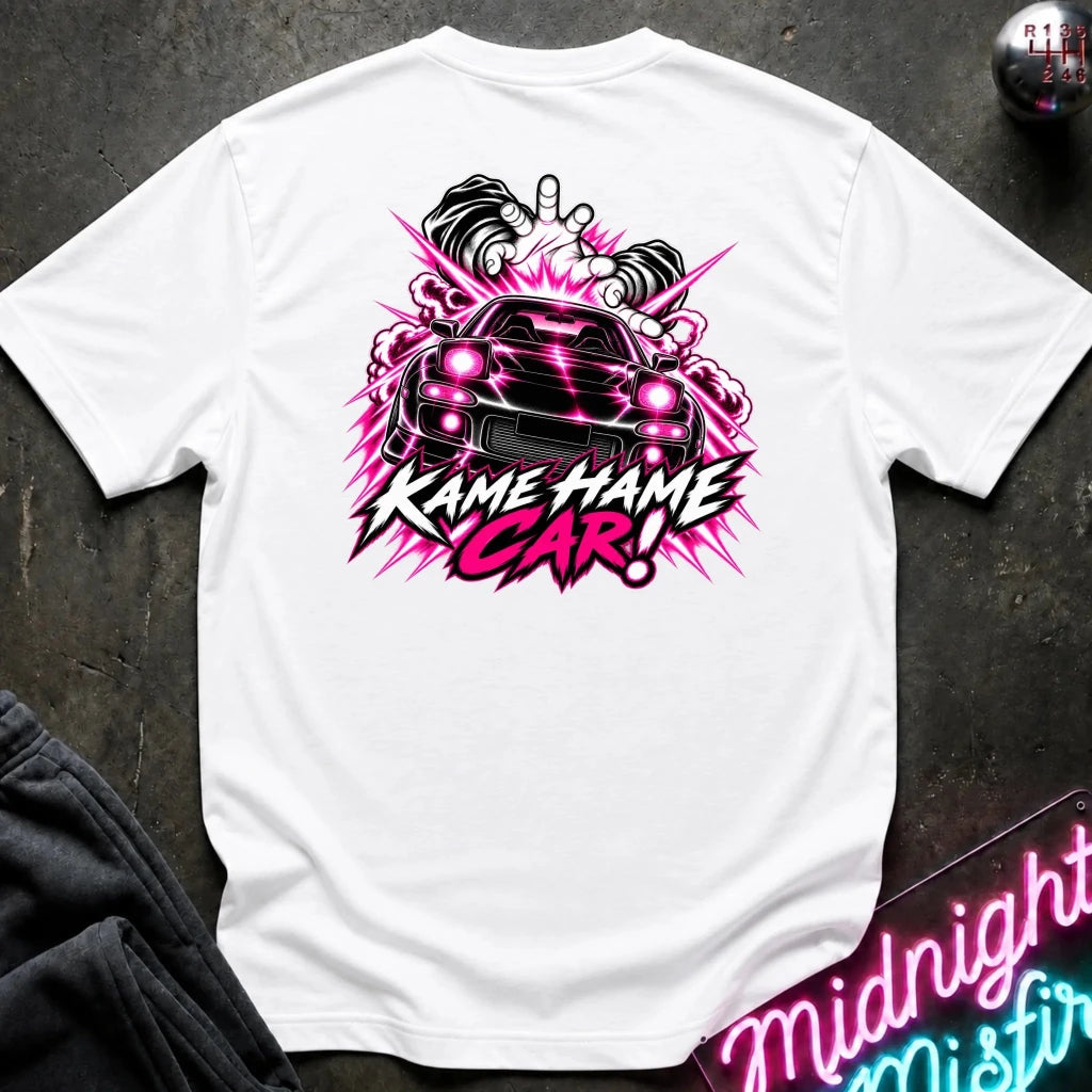 A white short-sleeve t-shirt laid flat on a dark textured surface, featuring a large graphic print of a black car with pink headlights and the text 'KAME HAME CAR!' in pink and black. 