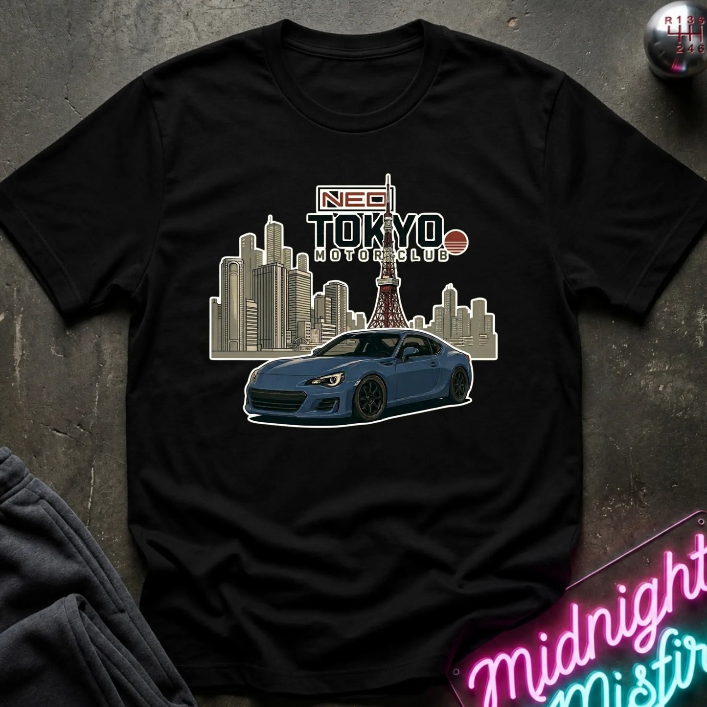 A black short-sleeve graphic T-shirt laid flat on a dark textured surface. The shirt features a printed design of a blue sports car in front of a city skyline and Tokyo Tower, with the text 'NEO TOKYO MOTOR CLUB' above it.