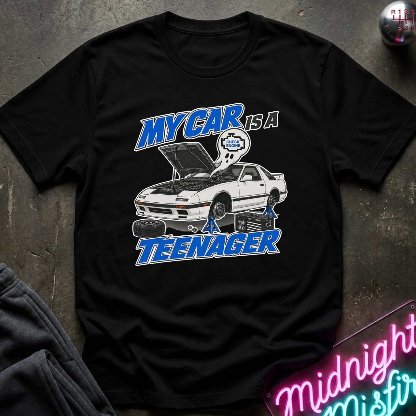 A black short-sleeve graphic t-shirt laid flat on a dark textured surface. The shirt features a graphic of a white sports car with its hood open, blue and white text reading 'MY CAR IS A TEENAGER'.