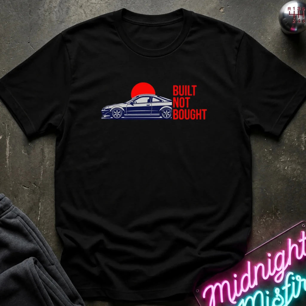A black short-sleeve graphic t-shirt laid flat on a dark textured surface. The shirt features a graphic of a blue sports car in front of a red sun with the text 'BUILT NOT BOUGHT' in red.