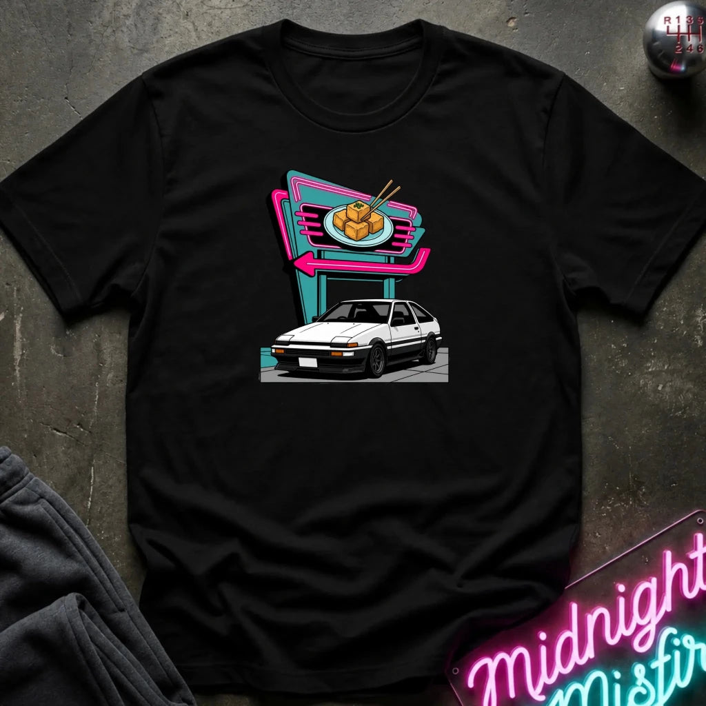 A black short-sleeve t-shirt laid flat on a dark textured surface, featuring a colorful graphic print of a white hatchback parked in front of a neon-style food sign. 
