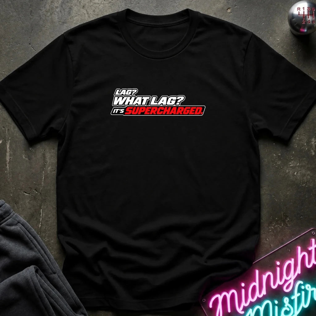 A black short-sleeve t-shirt with a graphic text print reading 'LAG? WHAT LAG? IT'S SUPERCHARGED' in white and red, laid flat on a dark textured surface with a neon sign and clothing prop nearby.