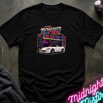 A black short-sleeve graphic T-shirt laid flat on a dark textured surface. The shirt features a colorful front print with the words 'Neo Tokyo Tire' above an illustration of a white convertible car in front of a neon-style shop sign.
