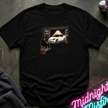A black short-sleeve graphic t-shirt laid flat on a dark textured surface. The graphic shows a white sports car in a garage with a person sitting in a chair under a hanging lamp.