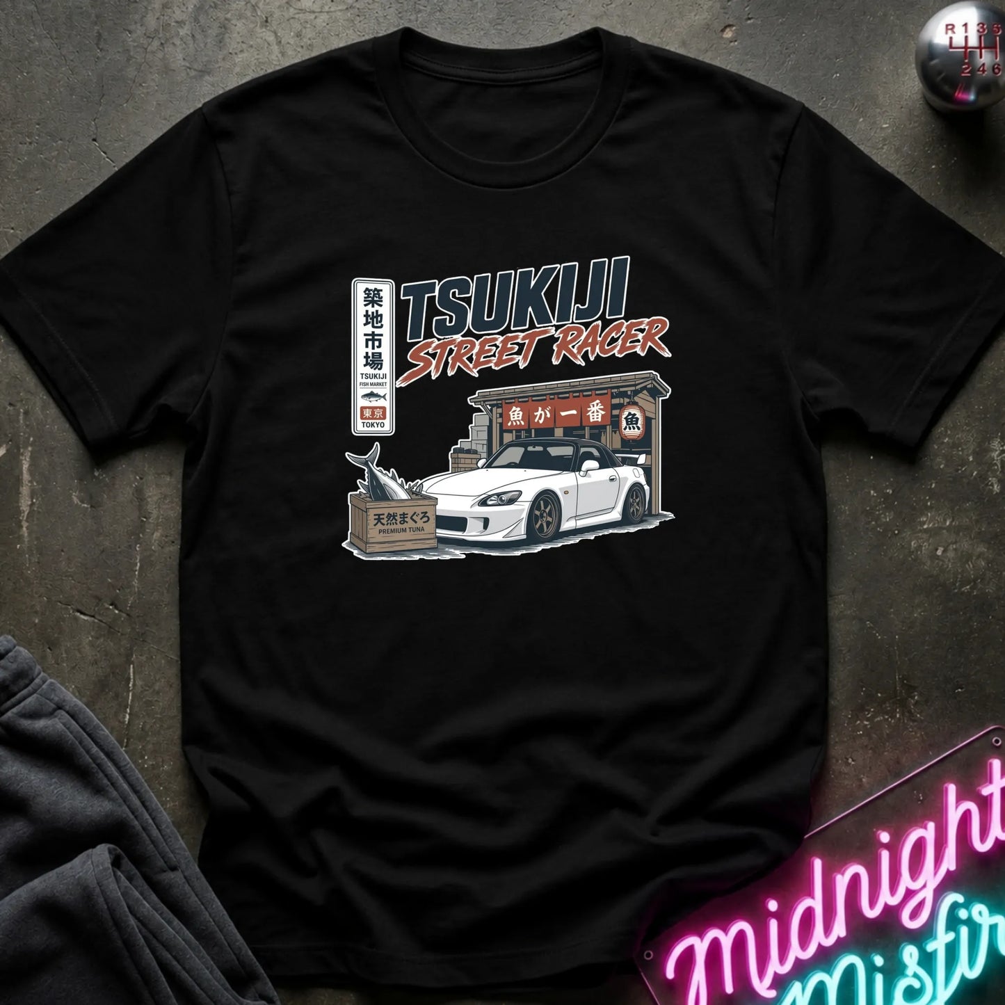 Black short-sleeve graphic T-shirt laid flat on and surrounding an illustration of a white sports car parked outside a fish market.