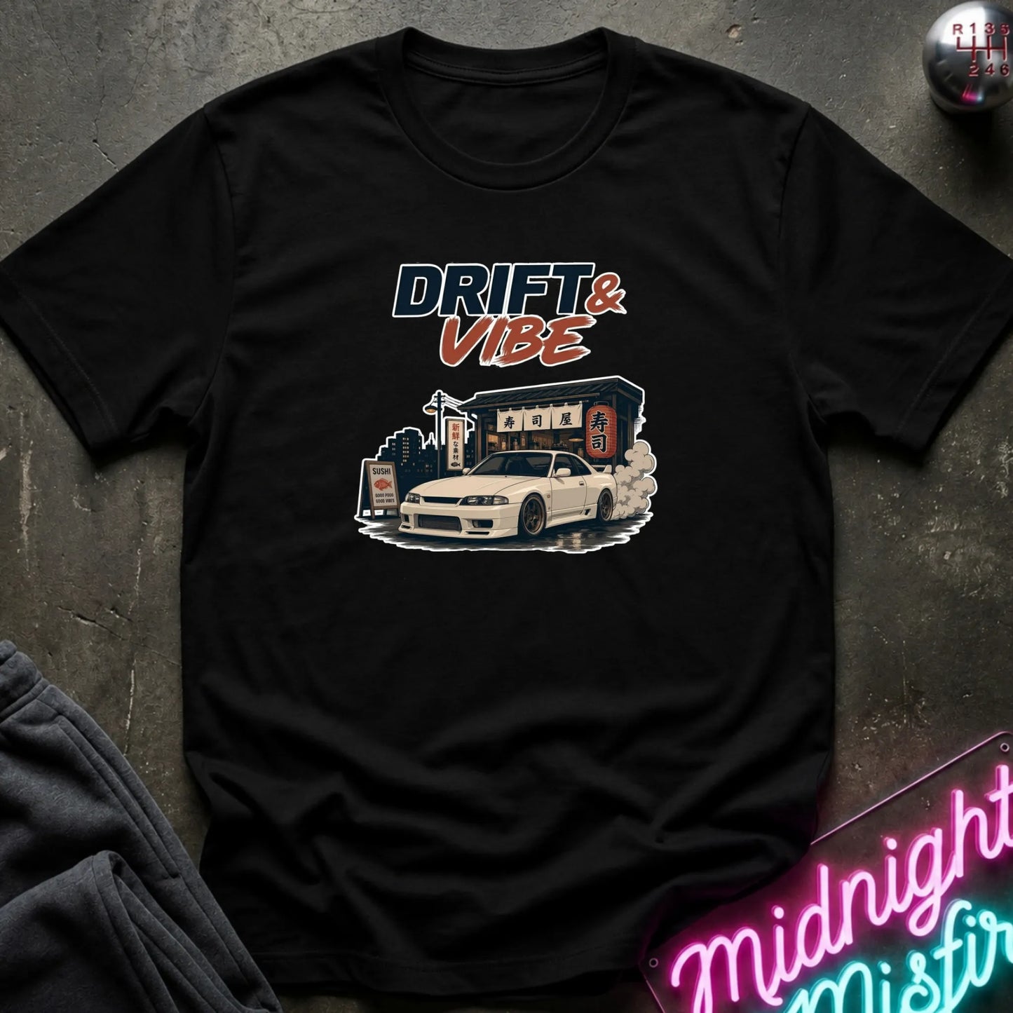 A black short-sleeve graphic t-shirt laid flat on a dark textured surface. The shirt features a graphic print with the text 'DRIFT & VIBE' in stylized blue and orange lettering above an illustration of a white sports car drifting in front of a building.
