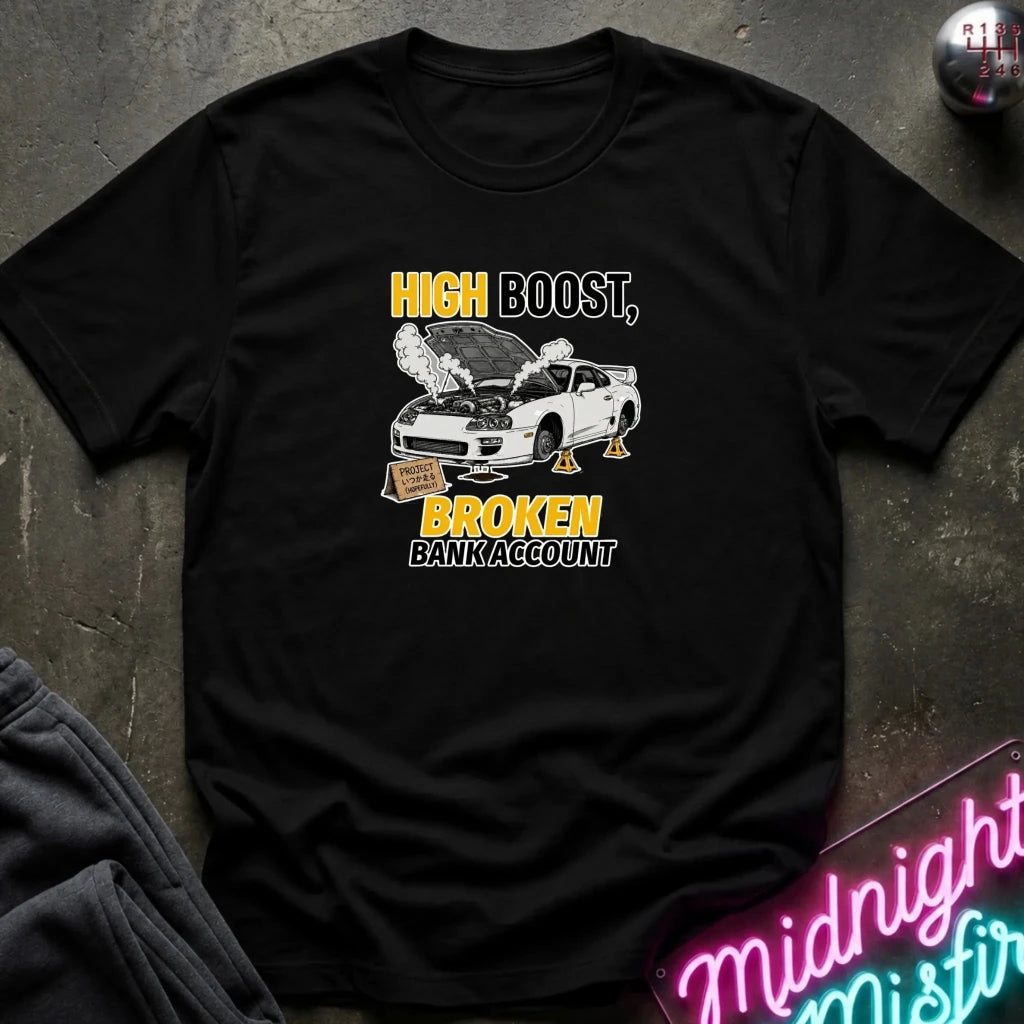 A black short-sleeve graphic t-shirt laid flat on a dark textured surface. The graphic features a white sports car with its hood open, emitting smoke, with yellow and white text reading 'HIGH BOOST, BROKEN BANK ACCOUNT'.