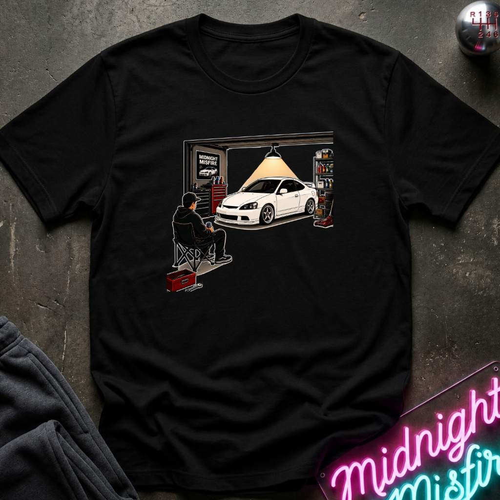 A black short-sleeve graphic t-shirt laid flat on a dark textured surface. The graphic shows a white sports car parked inside a garage with a person sitting in a chair under a hanging lamp.