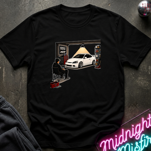 A black short-sleeve graphic t-shirt laid flat on a dark textured surface. The graphic shows a white sports car parked inside a garage with a person sitting in a chair under a hanging lamp.