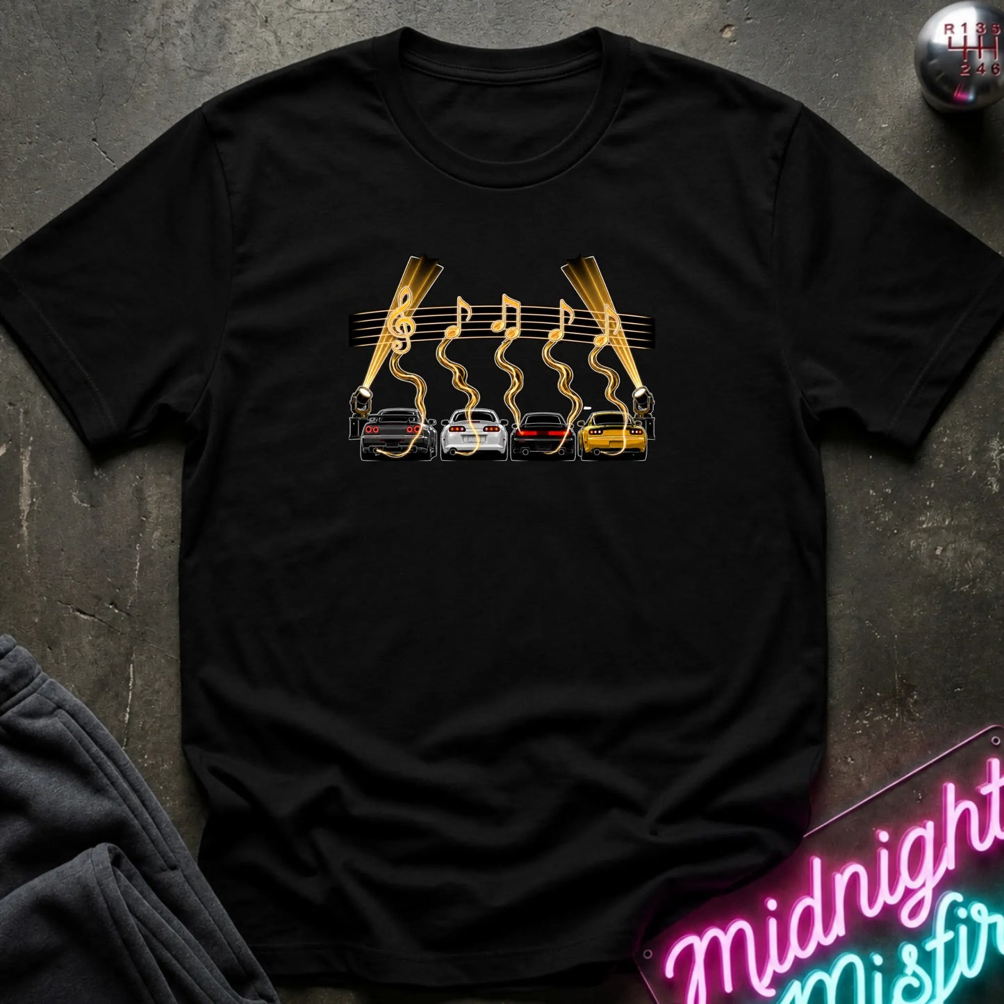 A black short-sleeve t-shirt laid flat on a dark textured surface, featuring a graphic print of four cars (red, white, gray, yellow) with gold wavy lines and musical notes above them, flanked by two  lights.